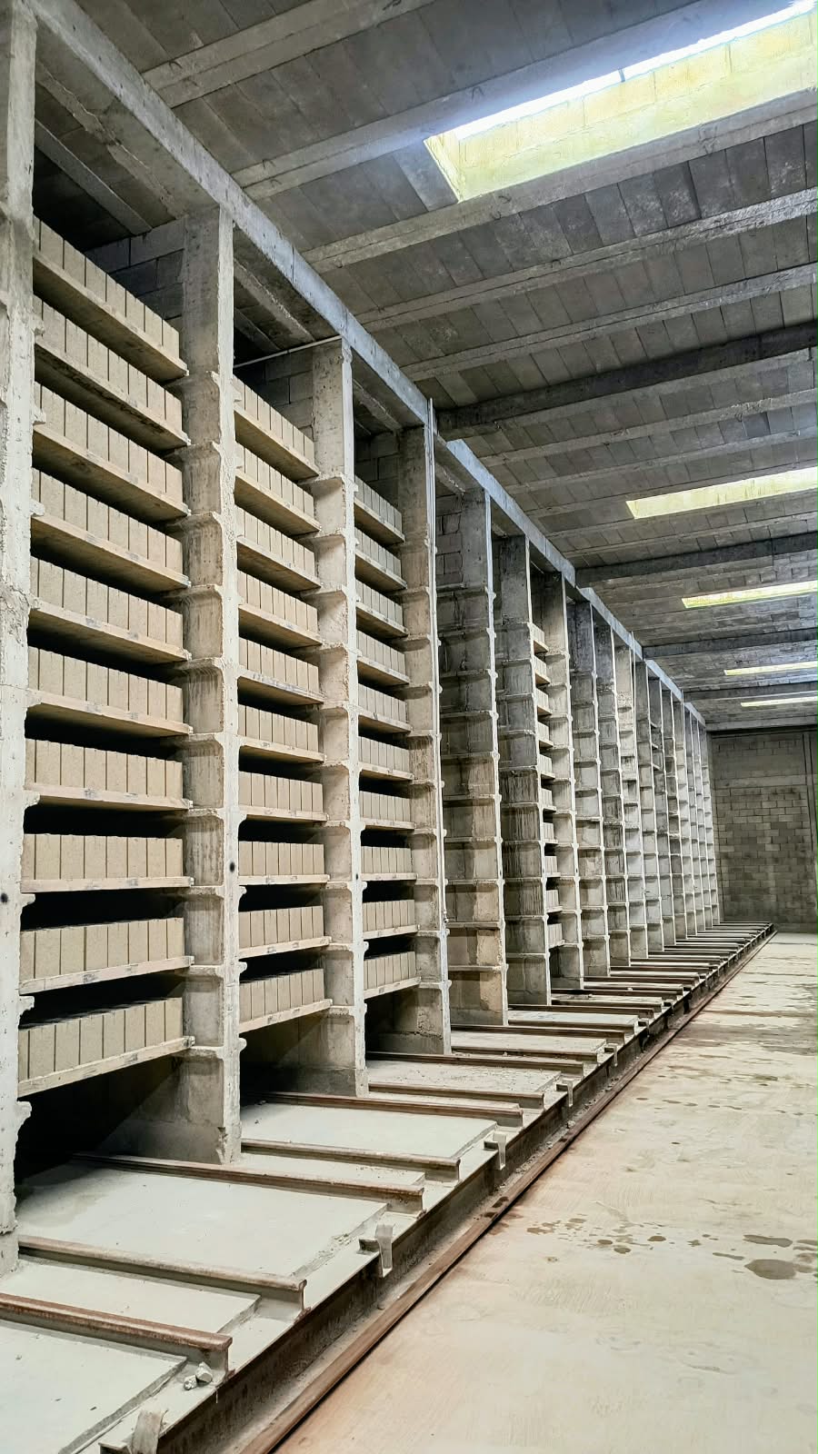 Nave de curado con racks de estantería de concreto cargados de bandejas con bloques recién formados, y rieles en el piso para el carro de transferencia